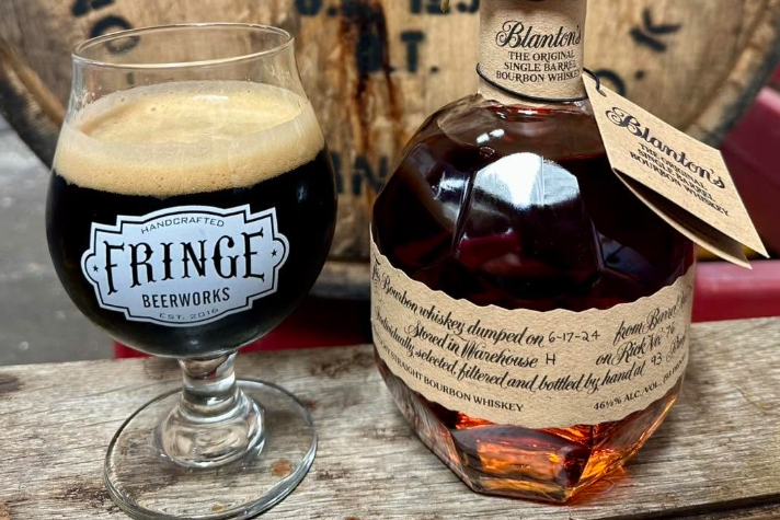 glass of a dark beer with a fringe beerworks logo and a bottle of whiskey